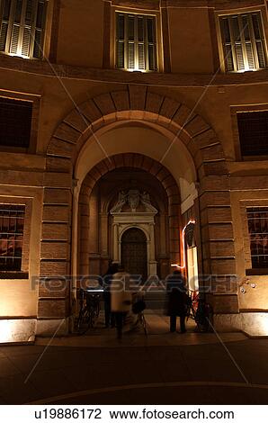 Stock Image -  Ferrara Theatre inner courtyard, illuminated circular building, with people walking with bicycles in shadows. Fotosearch