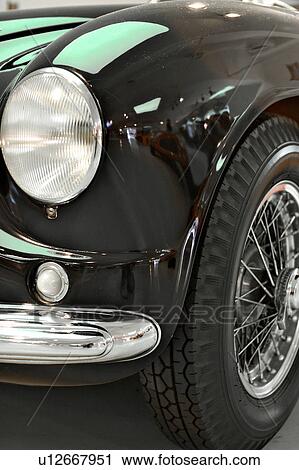 Stock Image - front quarter detail view of black Ferrari vintage model, showing bumper, headlamp, hood and wheel under green lighting in showroom. Fotosearch