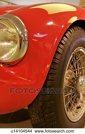 Picture - front quarter detail view of Ferrari vintage model, showing bumper, headlamp, hood and wheel under yellow lighting in showroom. Fotosearch