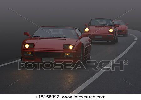 Stock Image - Front view of 2 red Ferraris and a Porsche coming up road in fog, with headlights on. Fotosearch