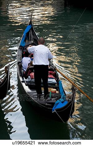 Stock Image - Gondola on canal, with back view of gondolier rowing, reflections in water. Fotosearch
