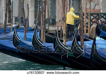 Stock Photo - Gondolas moored at canal side in winter rain with blue covers and a gondolier in a yellow jacket. Fotosearch
