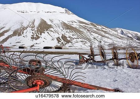 Stock Photo - hay making tools left for the winter in meadow of Piano Grande, Umbria, with snow in background on mountain slopes, and piles of wood in distance. Fotosearch
