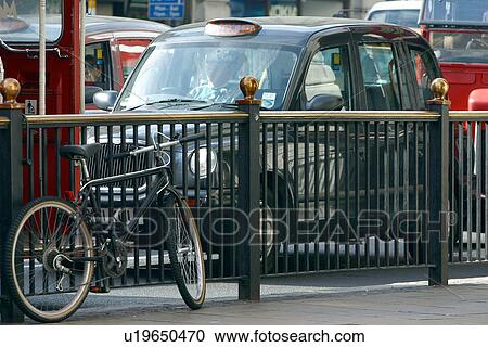 Stock Image - London taxi cab on street, beside iron railing, with parked bicycle and red double decker bus, London, England. Fotosearch