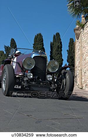 Low front-side view of burgundy antique Lagonda convertible, showing head lamps, grille and wheels, on road in Assisi, at Mille Miglia, with blue sky and cypress trees in background View Large Photo Image Stock Photo - Low front-side view of burgundy antique Lagonda convertible, showing head lamps, grille and wheels, on road in Assisi, at Mille Miglia, with blue sky and cypress trees in background. Fotosearch