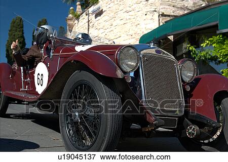 Low front-side view of burgundy antique car with driver and passenger, showing head lamps, grille and wheels, on road in Assisi, at Mille Miglia, with blue sky and cypress trees in background View Large Photo Image Stock Photo - Low front-side view of burgundy antique car with driver and passenger, showing head lamps, grille and wheels, on road in Assisi, at Mille Miglia, with blue sky and cypress trees in background. Fotosearch