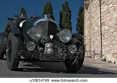 Low front-side view of green antique Bentley with driver and passenger, showing head lamps, grille and wheels, on road in Assisi, at Mille Miglia, with blue sky and cypress trees in background View Large Photo Image Stock Photo - Low front-side view of green antique Bentley with driver and passenger, showing head lamps, grille and wheels, on road in Assisi, at Mille Miglia, with blue sky and cypress trees in background. Fotosearch