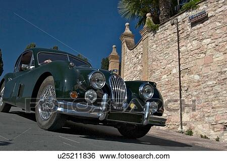 Stock Photograph - Low front-side view of green Jaguar antique car showing head lamps, grille and wheels, on road in Assisi, at Mille Miglia, with blue sky and trees in background. Fotosearch