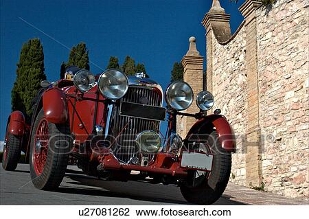 Low front-side view of red Lagonda antique car with driver, showing head lamps, grille and wheels, on road in Assisi, at Mille Miglia, with blue sky and cypress trees in background View Large Photo Image Stock Image - Low front-side view of red Lagonda antique car with driver, showing head lamps, grille and wheels, on road in Assisi, at Mille Miglia, with blue sky and cypress trees in background. Fotosearch