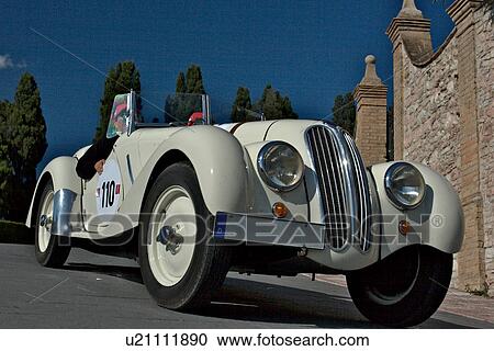 Low front-side view of white antique BMW with driver and passenger, showing head lamps, grille and wheels, on road in Assisi, at Mille Miglia, with blue sky and cypress trees in background View Large Photo Image Stock Image - Low front-side view of white antique BMW with driver and passenger, showing head lamps, grille and wheels, on road in Assisi, at Mille Miglia, with blue sky and cypress trees in background. Fotosearch