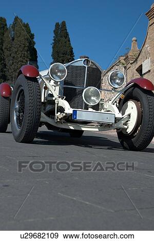 Stock Photo - Low front view of white antique Mercedes with red trim on wheel arches, showing head lamps, grille and wheels, on road in Assisi, at Mille Miglia, with blue sky and cypress trees in background. Fotosearch