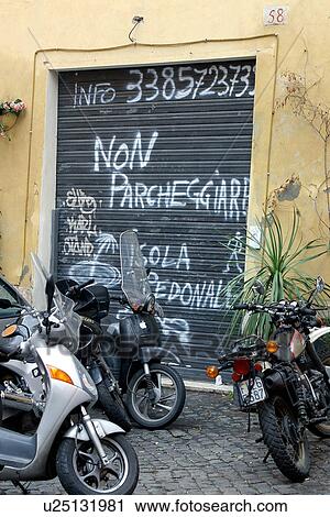 Stock Image - Motor bikes parked outside delapidated garage with "no parking" signs scrawled on metal door, Rome, Italy. Fotosearch