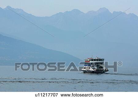 Stock Photo - Passenger ferry boat in motion across Lake Como, Italy, in morning sunlight, with blue water, mountain range, and sky. Fotosearch