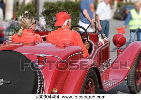Picture - Rear quarter view of antique red car with driver and passenger in red outfits, showing side of car with running board. Fotosearch