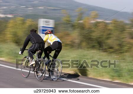 Stock Image - Rear view of 2 cyclists in training gear, cycling along road in morning autumn sun, Umbria, Italy, with green fields and trees in background. Fotosearch