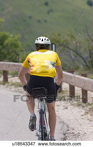 Stock Photo - Rear view of cyclist in yellow shirt, black shorts, cycling up mountain on winding road in morning sun, Umbria, Italy, with green fields and trees in background. Fotosearch