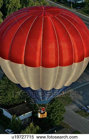 Stock Photography - Red white and blue hot air balloon, seen from above, flying over grass and trees at dusk. Fotosearch