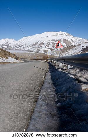 Stock Photo - Road curving, and road sign at Piano Grande, Umbria, with snow on mountains, brilliant sunshine and blue sky on cloudless winter day. Fotosearch