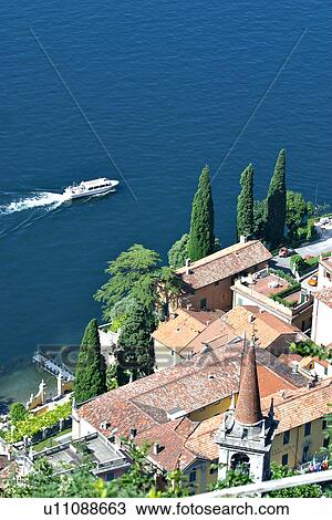 Rooftop view of lakeside village of Castello di Vezio buildings with church bell tower, in bright morning sunshine, with boat in motion in blue water of Lake Como View Large Photo Image Stock Image - Rooftop view of lakeside village of Castello di Vezio buildings with church bell tower, in bright morning sunshine, with boat in motion in blue water of Lake Como. Fotosearch