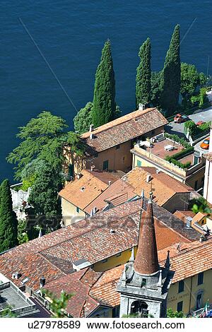 Stock Photo - Rooftop view of lakeside village of Castello di Vezio buildings with church bell tower, in bright morning sunshine, and blue water of Lake Como in background. Fotosearch