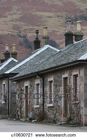 Stock Image - Row of Highland cottages, granite block construction, slate tile roofs with lead trim, chimneys against pink and yellow background of heather covered hills, Scotland. Fotosearch