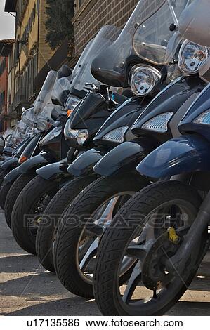 Stock Photograph - Row of motorbikes parked on street in front of Renaissance building, showing front wheels, windscreen, headlamps and cowling detail, Florence, Italy. Fotosearch