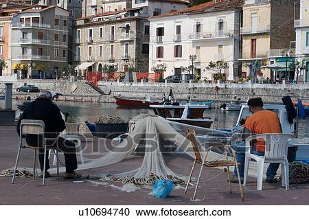 Stock Image - Scario harbour with boats, fisherman repairing nets, small fishing boats moored, and town of Scario beyond, on sunny, summer morning. Fotosearch