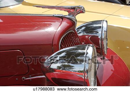 Stock Image - Side view detail of antique red Citroen hood ornament and silver head lamps, with tree reflection and yellow car in background. Fotosearch