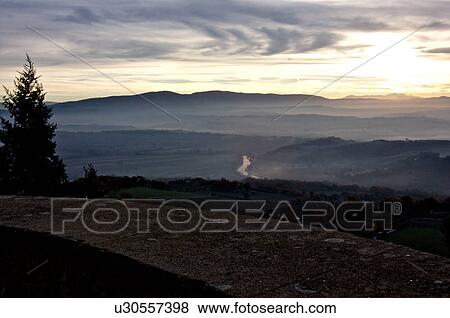 Stock Photo - Sunrise over Tiber valley with grey and golden sky in the early morning mist of winter, with tree outline and town stone wall in foreground, and River Tiber curving beside hill. Fotosearch