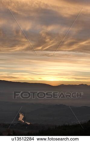 Picture - Sunrise over Tiber valley with grey pink and golden sky in the early morning mist of winter, with tree outline in foreground, and River Tiber curving beside hill. Fotosearch