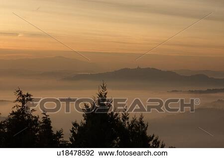 Stock Image - Sunrise over Tiber valley with grey pink and golden sky in the early morning mist of winter, with tree outline in foreground, and hilltop town of Todi in distance. Fotosearch
