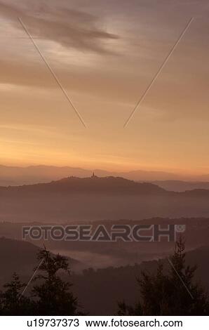 Stock Image - Sunrise over Tiber valley with grey pink and golden sky in the early morning mist of winter, with tree outline in foreground, and hilltop town of Todi in distance. Fotosearch