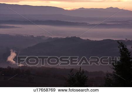 Stock Photo - Sunrise over Tiber valley with grey pink and golden sky in the early morning mist of winter, with tree outline in foreground, and River Tiber curving beside hill. Fotosearch