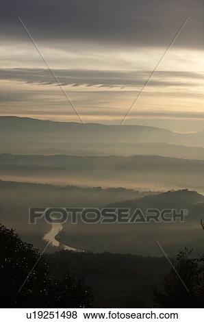Stock Photo - Sunrise over Tiber valley with grey pink and golden sky in the early morning mist of winter, with tree outline in foreground, and River Tiber curving beside hill. Fotosearch