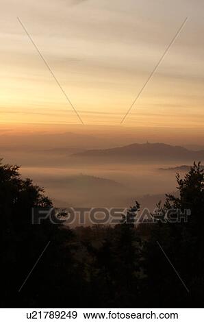 Stock Photo - Sunrise over Tiber valley with grey pink and golden sky in the early morning mist of winter, with tree outline in foreground, and hilltop town of Todi in distance. Fotosearch