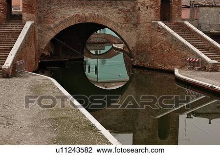 View of 17th century Trepponte bridge, Comacchio, on winter afternoon, showing pavement alongside canals, arches spanning the water, towers, and reflections of pedestrians crossing View Large Photo Image Stock Image - View of 17th century Trepponte bridge, Comacchio, on winter afternoon, showing pavement alongside canals, arches spanning the water, towers, and reflections of pedestrians crossing. Fotosearch