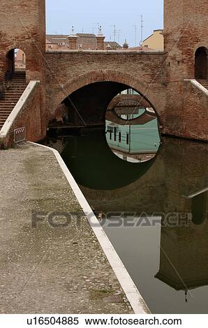Stock Photography - View of 17th century Trepponte bridge, Comacchio, on winter afternoon, showing pavement alongside canals, arches spanning the water, towers, and reflections of pedestrians crossing. Fotosearch