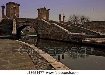 Stock Photo - View of 17th century Trepponte red brick bridge, Comacchio, on winter afternoon, showing pavement alongside canals, arches spanning the water, towers, and reflections of pedestrians crossing. Fotosearch