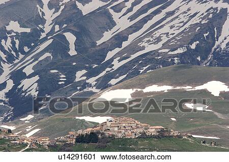 Stock Image - View of Castellucio, Piano Grande, as the snows melt on the mountains behind the town, forming abstract patterns as a background to the dwarfed town. Fotosearch
