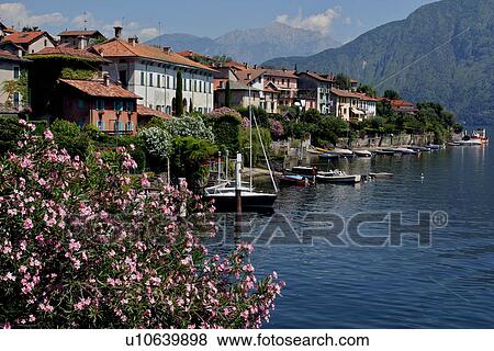 Stock Photo - View of Lake Como village with boats moored, flowers in foreground, houses and mountains in background. Fotosearch