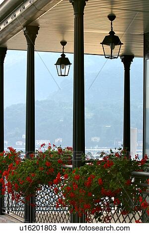 Stock Image - View of lakeside restaurant covered outdoor terrace, with mesh iron railing, black columns, ornamental lamps hanging, and baskets of red geraniums, with lake and mountains beyond, Lake Como, Italy. Fotosearch