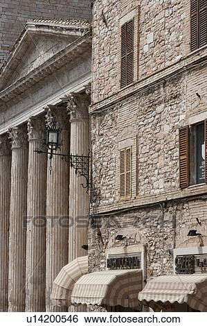 Stock Photograph - View of Roman Temple of Minerva in Piazza del Comune, Assisi, with adjacent stone facade and canopies of shop fronts matching colour of light beige. Fotosearch