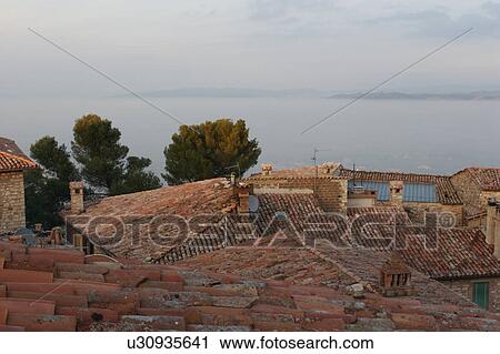 Stock Image - View of roof tops of Umbrian medieval village with dense valley fog creating appearance of a lake in the valley beyond, and mountains in the distance. Fotosearch