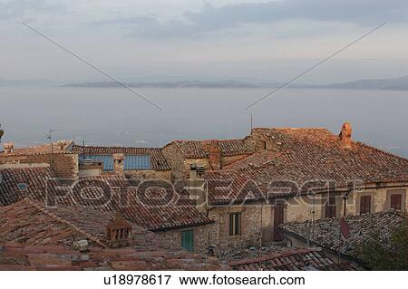 Stock Photo - View of roof tops of Umbrian medieval village with dense valley fog creating appearance of a lake in the valley beyond, and mountains in the distance. Fotosearch