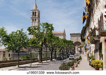Stock Photo - View of street in Assisi, leading to Church of Santa Chiara, with bell tower and medieval gate of city wall, cars, flags, trees and flowers on sunny summer day with blue sky. Fotosearch