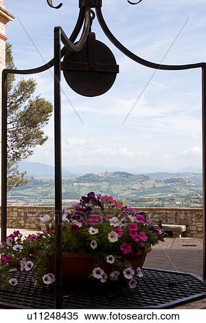 Stock Photography - View of Tiber valley with hilltop town of Todi seen beyond medieval well, with wrought iron detail, and flowers, blue sky on sunny summer afternoon. Fotosearch