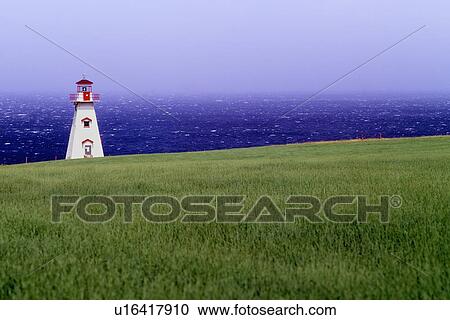 Stock Image - White traditional Canadian lighthouse with red detail on edge of grassy cliff, against blue sky, with dark blue sea and waves beyond. Fotosearch
