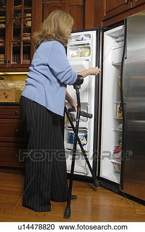 Stock Image - Female with arm crutches looks in refrigerator.. Fotosearch