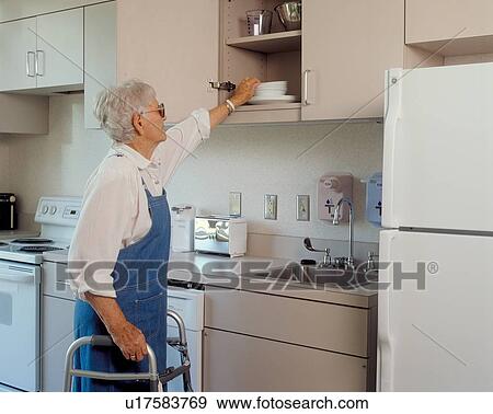 Stock Photo - Older woman with a disability, utilizing a walker to assist with her mobility, getting a dish from her cabinet to prepare a meal.. Fotosearch