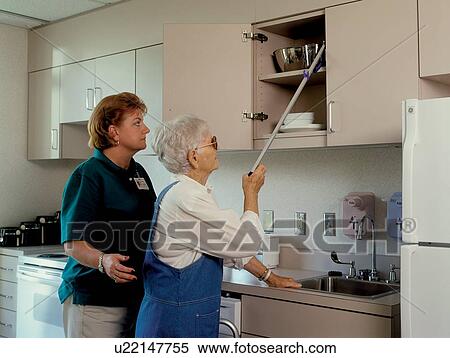 Stock Photography - Older woman with a disability utilizing a reaching device or reacher to retrieve a dish from the top cabinet. An assistant is standing by to offer assistance.. Fotosearch